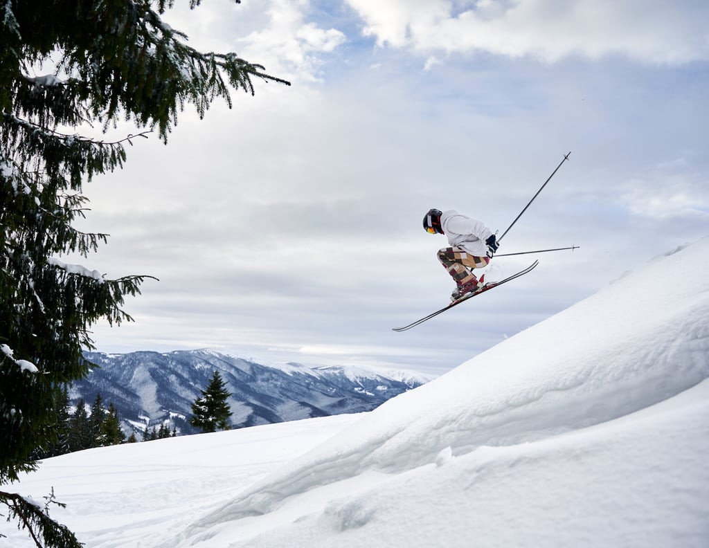 Side view of male skier making jump while sliding down snow-covered slope on skis under beautiful cloudy sky. Man freerider in helmet skiing on fresh powder snow in mountains.