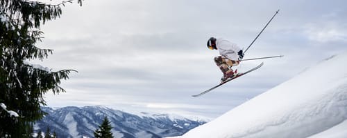 Side view of male skier making jump while sliding down snow-covered slope on skis under beautiful cloudy sky. Man freerider in helmet skiing on fresh powder snow in mountains.