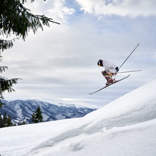 Side view of male skier making jump while sliding down snow-covered slope on skis under beautiful cloudy sky. Man freerider in helmet skiing on fresh powder snow in mountains.